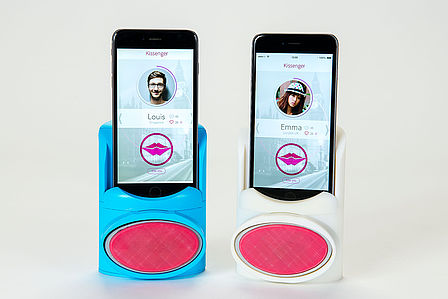 Ein blauer und weisser, barrenförmiger "Kissenger". Darin stecken jeweils ein Smartphone mit der Kissenger-App am Screen. Jeder Kissenger besitzt euine rote, ovale Fläche zu Küssen.