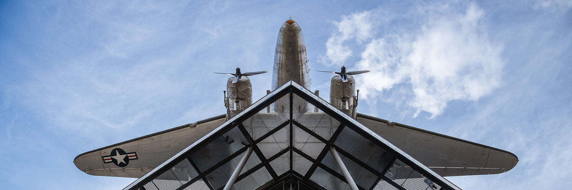 Blick von unten auf einen Neubau mit Glasfassade und Stahlstreben. Darüber hängt ein Flugzeug.