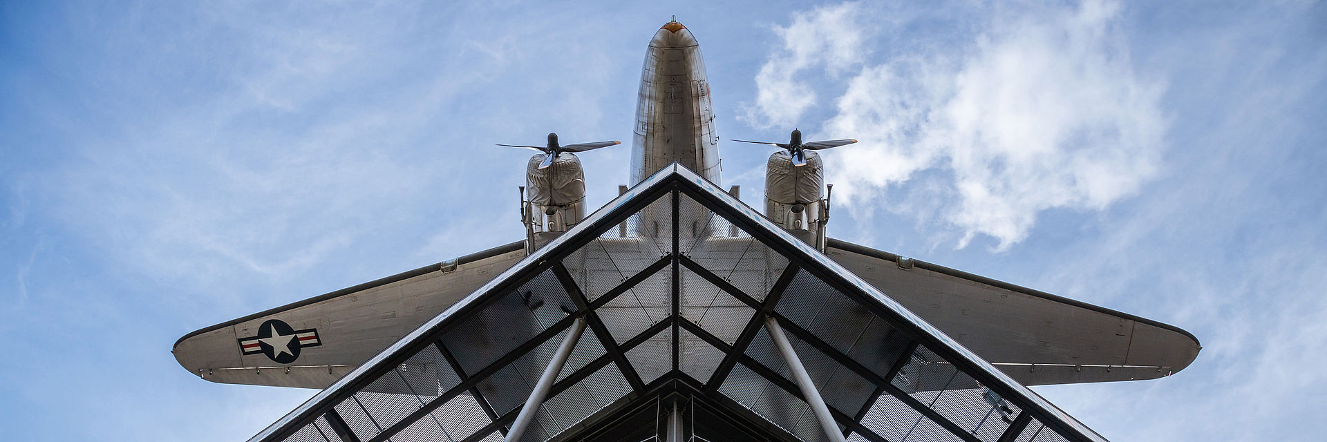 Blick von unten auf einen Neubau mit Glasfassade und Stahlstreben. Darüber hängt ein Flugzeug.