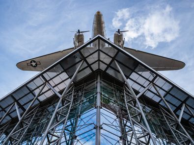 Blick von unten auf einen Neubau mit Glasfassade und Stahlstreben. Darüber hängt ein Flugzeug.
