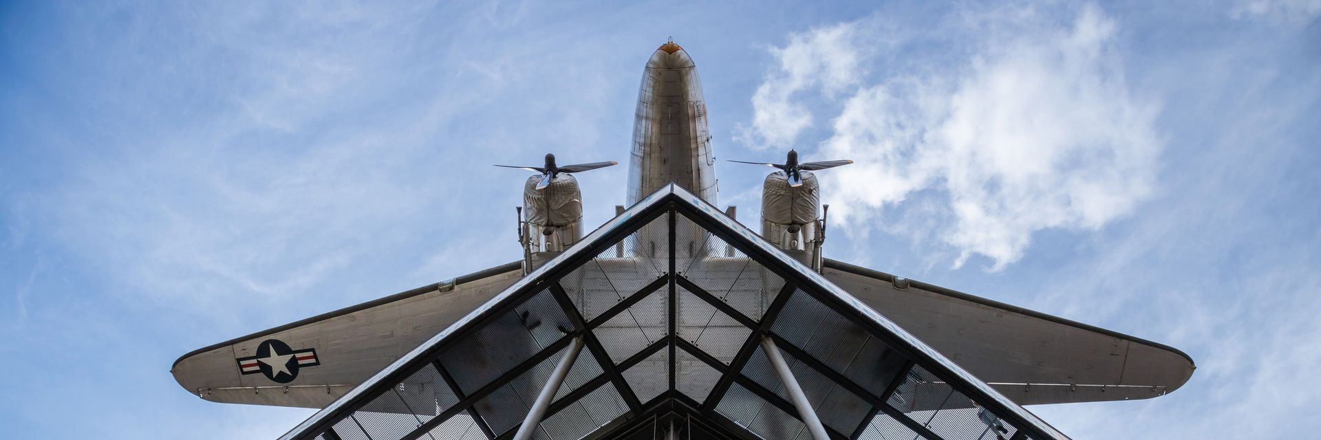 Blick von unten auf einen Neubau mit Glasfassade und Stahlstreben. Darüber hängt ein Flugzeug.