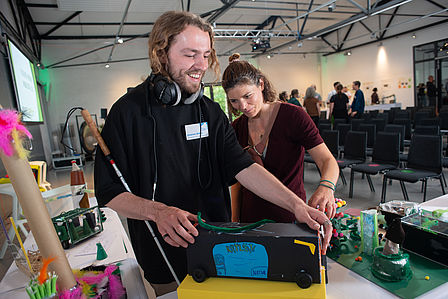 Zwei Menschen stehen im Konferenzraum. Sie ertasten lächelnd die gebastelten bunten Modelle der SchülerInnen. Am Arm der linken Person lehnt ein Blindenstock. 