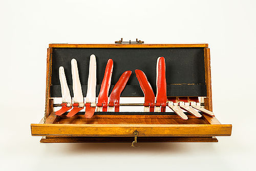 The finger calculator consists of a brown wooden case, the front part of which can be opened downwards to form a kind of showcase during lessons. The outlines of the ten metal fingers of two "hands" are displayed. There is a set of duplicate fingers hidden behind the visible ones. There are thus two rows of fingers. The front row is made up of red metal fingers behind which are identically shaped white ones. Both rows of fingers can be folded down towards the front by means of a simple mechanism. 