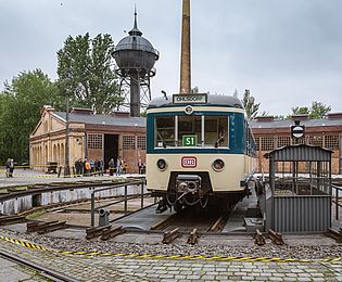 Blick auf die Drehscheibe am Lokschuppen im Museumspark. Auf der Drehscheibe steht eine blau-beige S-Bahn. Im Hintergrund sind ein metallener Wasserturm und ein Schornstein zu sehen.