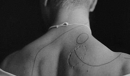 Das Schwarz-Weiß-Foto zeigt Nacken und Rücken einer Person mit kurzem Haar. Die leicht herabgerutschte Bluse gibt die Schulterpartie frei. Auf der Haut sieht man handgezeichnete Tattoos aus drei feinen teilweise gepunkteten Linien und einem Kreis. Auf dem rechten Schulterblatt ist eine narbenähnliche Kreisstruktur zu sehen, die wie eine Bissspur aussieht.