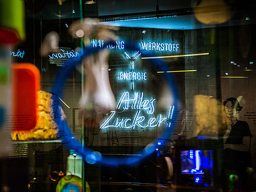 Blick durch eine Vitrine auf ein großes Neonschild. Das Schild besteht aus dem Schriftzug „Alles Zucker!“. Darüber befinden sich die Worte „Werkstoff“, „Nahrung“ und „Energie“, die mit Strichen miteinander verbunden sind. Die Ränder der Aufnahme sind unscharf.