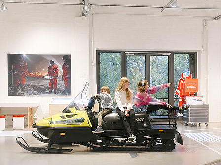 Drei Kinder sitzen auf einem gel-schwarzen Schneemobil in einer Ausstellungshalle.