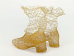 Zwei Stiefel stehen nebeneinander. Sie sind aus feinem Kupferdraht gewebt, der wie ein feiner Goldfaden aussieht. An den Füßen und den Hacken ist das Material dichter gewoben. Oben wird das Material luftiger, als würden die Stiefel sich auflösen.