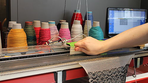 Eine Person bedient eine Strickmaschine, die auf einem Tisch steht. Ein schwarz-weiß gemusterter Stoff entsteht durch das Hin- und Herbewegen des Schlittens. Im Hintergrund stehen Spulen mit bunten Stofffäden, daneben ein aufgeklappter Laptop.