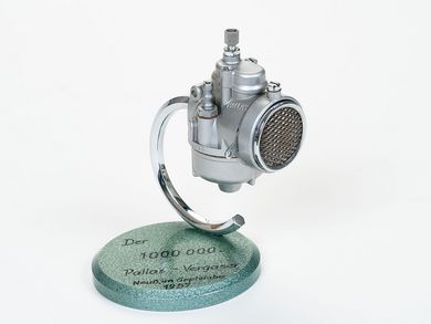 Auf einer runden Grundplatte ist der millionste Vergaser der Marke Pallas montiert. Es handelt sich um einen kleinen Moped-Vergaser. Die Grundplatte ist mit grüner Hammerschlaglackierung versehen, auf der das Jubiläum und das Datum in schwarzer Schrift notiert wurden.
