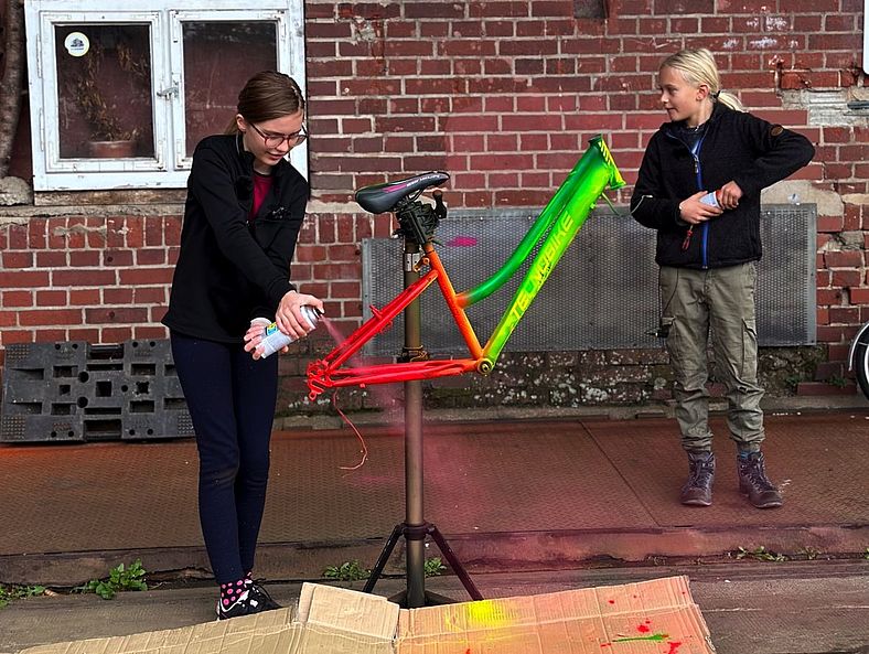 Zwei Kinder sprühen bunte Farbe auf den Rahmen eines Fahrrads, das auf einem Ständer montiert ist. Im Hintergrund ist eine Backsteinwand mit Schildern und alten Gerätschaften zu sehen.