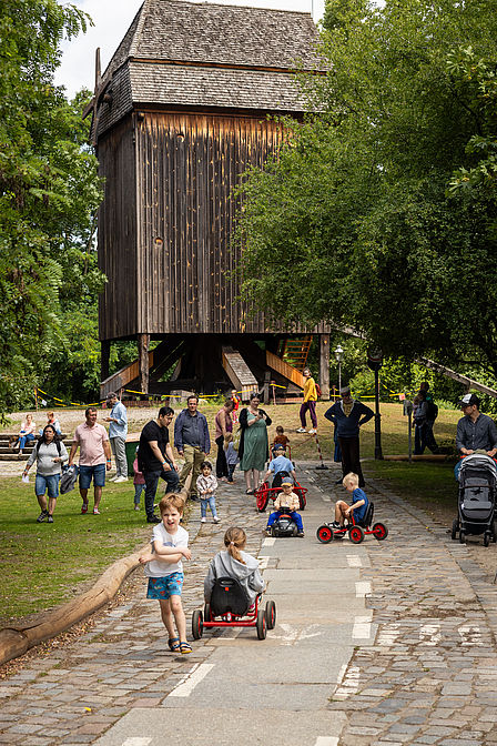 Vor einer Bockwindmühle fahren Kinder auf fantasievollen Gefährten.