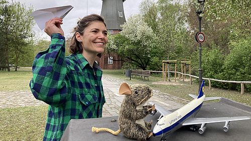Im Museumspark. Im Hintergrund die Holländermühle. Im Vordergrund sitzt "Nanu" die Maus neben einem Modellflugzeug. Links neben ihr wirft eine Museumpädagoging ein Papierflugzeug. 