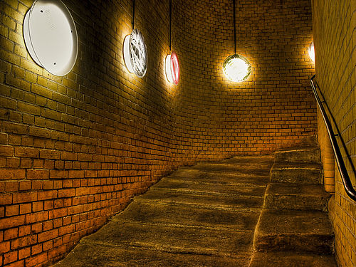 Das Treppenhaus einer Pferdewendeltreppe. Die Wand des Treppenhauses besteht aus braunen Klinkersteinen. Daran sind runde, flache Lampen angebracht. Die Treppe ist zweigeteilt. Außen verlaufen breite, tiefe ausgetretene Stufen, innen sind sie kürzer und weniger ausgetreten.
