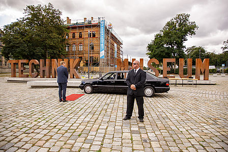 Schwarze Limousine auf dem Vorplatz, Schriftzug "Technik Museum" im Hintergrund, zwei Männer in schwarzen Anzügen.
