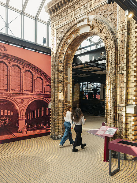 Zwei Frauen laufen durch das originale Fürstenportal des Anhalter Bahnhofs in Richtung der Ausstellung Schienenverkehr. Durch das Portal kann man im Hintergrund eine Eisenbahn erkennen.