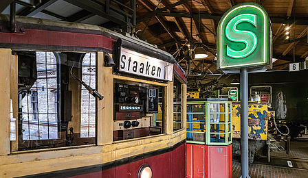 Der rot-gelbe Führerstand eines S-Bahnwagens steht im Vordergrund, dahinter leuchtet ein großes S-Bahn Schild in der Ausstellung.