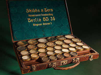 Lederkoffer mit verschiedenen kleinen Gefäßen, in denen sich Proben kolonialer Produkte wie Kakao befinden. Die Innenseite des Kofferdeckels trägt den Firmennamen Skibbe & Zorn.