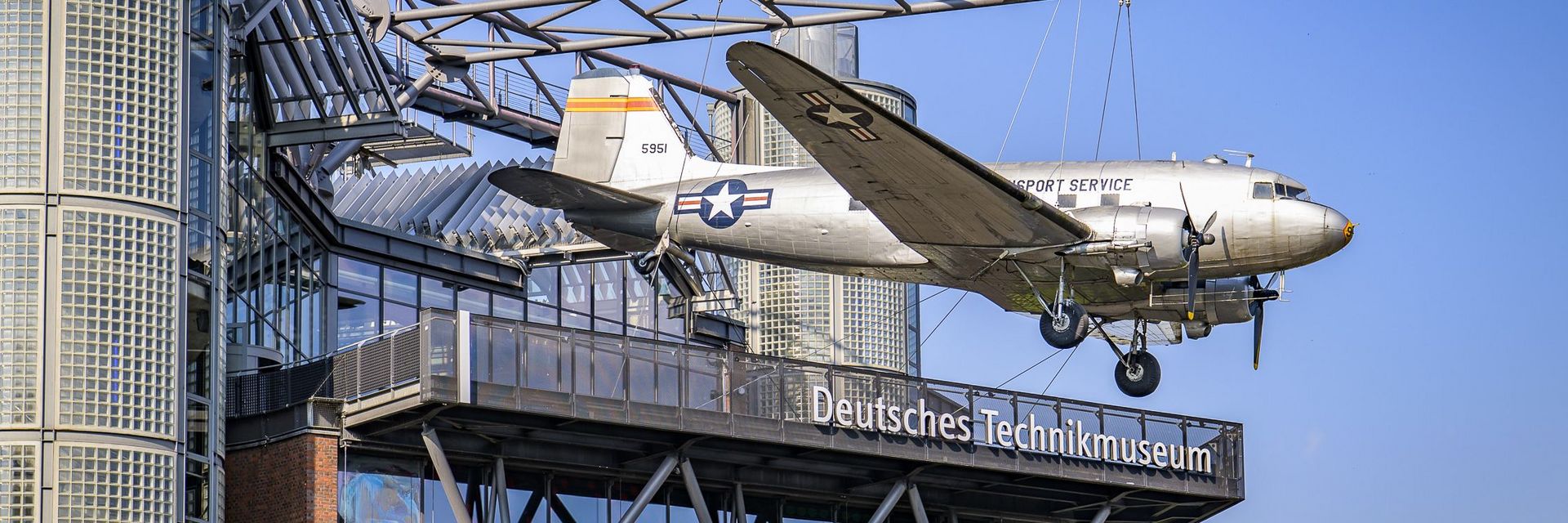 Ein Neubaugebäude mit Glasfassade. An einer Stahlkonstruktion auf dem Dach hängt ein Flugzeug.