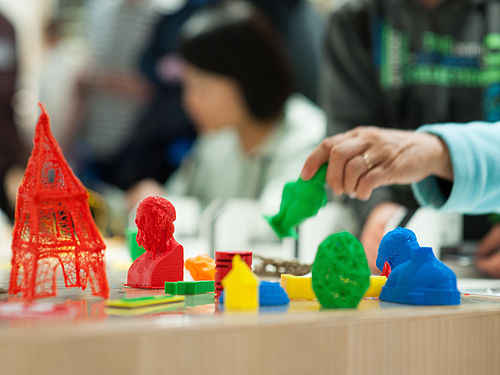 Auf einem Tisch stehen Figuren, Gegenstände und Kunstwerke, die mit einem 3D-Drucker aus verschiedenfarbigem, recyclebarem Plastik gedruckt wurden. Links steht eine rote, etwa 15 Zentimeter hohe, mit einem 3D-Stift gezeichnete, grobe Nachbildung des Eifelturms.