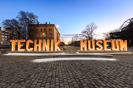 Auf einem gepflasterten Platz sieht man eine aus Metallbuchstaben erstellte Wortskulptur, die sich „Technik Museum“ liest. Im Hintergrund ein Backsteingebäude.