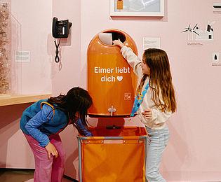 Ein orangener Mülleimer, auf dem „Eimer liebt Dich“ steht. Zwei Mädchen befüllen den Eimer, darunter eine orangefarbene Tasche, die den Müll beim Entleeren auffängt. Dahinter ein Display, das zeigt, wann der Mülleimer voll ist.