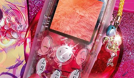 Ein transparentes Handy mit Anhänger liegt auf einem pinken und glitzerfarbenen Untergrund, darauf sind verschiedene Formen und weitere transparente Handygehäuse erkennbar.