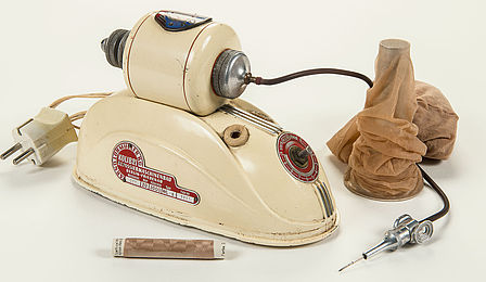 Zu sehen ist eine cremefarbige Maschine, die entfernt an ein Bügeleisen erinnert. Sie hat ein breites Standbein und einen Stecker, einen braunen Schlauch und eine silberfarbene Nadel. Daneben steht ein Ständer mit einer beige-braunen Feinstrumpfhose, davor liegt eine Garnrolle in der gleichen Farbe.