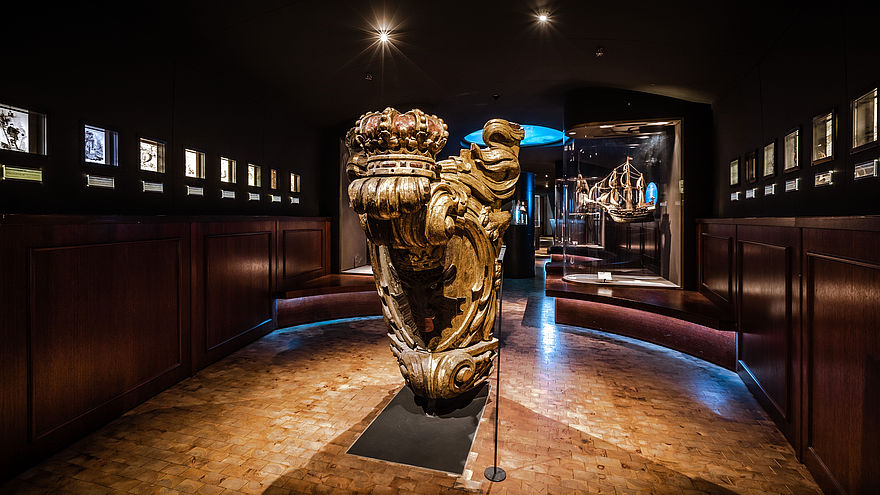 Blick in die Schifffahrt-Ausstellung: Zentral im Raum steht eine hölzerne Galionsfigur. An beiden Seiten verläuft ein Band aus kleinen Leuchtkästen. Im Hintergrund ist eine Vitrine mit einem Schiffsmodell zu sehen.
