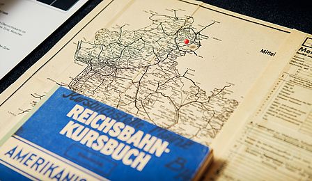 A map with railway routes; the cities of Frankfurt and Wiesbaden are on the map. Above it is a blue book with the inscription "Reichsbahn Kursbuch". 