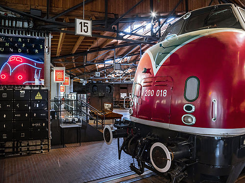 Die Schnellzuglokomotive V 200 steht in den historischen Lokschuppen des Deutschen Technikmuseums auf in den Holzboden eingelassenen Schienen. Ihr abgerundeter Rumpf ist Feuerrot lackiert. Links daneben befindet sich ein Neonschild in Form der Lokomotive. 
