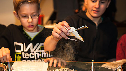Zwei Jungen sitzen an einem Tisch. Vor ihnen, auf einer dunklen Tischplatte, liegen ein Blatt Papier, zwei silberne, metallische Hütchen sowie eine kleine runde Korkplatte mit dunkler Oberfläche und Staubmuster. Der Junge rechts hält seine Hand mit einem hellen Stoffbeutel über die Korkplatte. Aus dem Beutel heraus staubt ein Pulver.