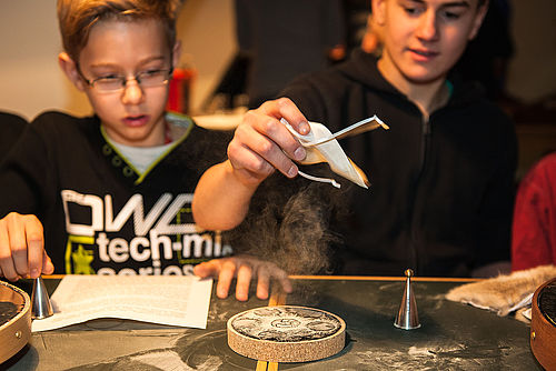 Zwei Jungen sitzen an einem Tisch. Vor ihnen, auf einer dunklen Tischplatte, liegen ein Blatt Papier, zwei silberne, metallische Hütchen sowie eine kleine runde Korkplatte mit dunkler Oberfläche und Staubmuster. Der Junge rechts hält seine Hand mit einem hellen Stoffbeutel über die Korkplatte. Aus dem Beutel heraus staubt ein Pulver.