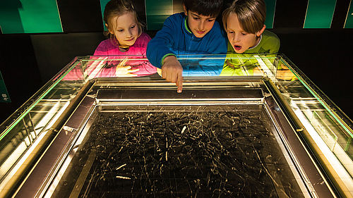 Unter einer am Rand beleuchteten Glasabdeckung befindet sich eine schwarze Fläche, auf der viele verschiedene kleine weiße nebelartige Streifen zu sehen sind. Drei Kinder schauen auf diese Streifen. Das Kind in der Mitte zeigt darauf.