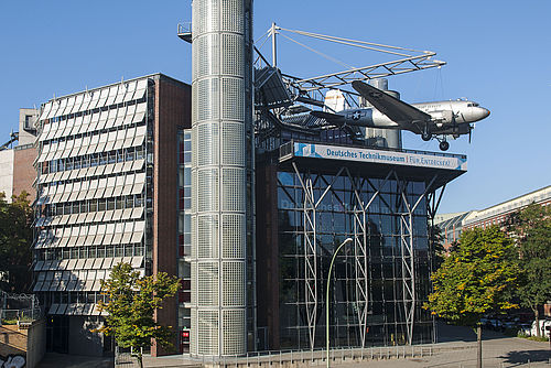 Ein modernes Neubaugebäude mit Glasfassade. An einer Stahlkonstruktion auf dem Dach hängt ein Flugzeug.