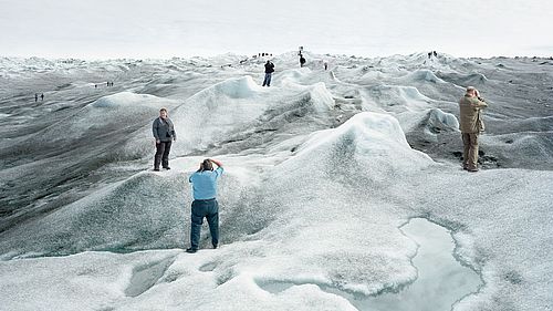 Eine Gruppe von Touristinnen und Touristen steht auf einer schmelzenden Eislandschaft und posiert an verschiedenen Punkten des Bildes für Fotos. An vielen Stellen der Eislandschaft hat sich schwarzer Ruß abgesetzt, das Eis ist dort sehr dunkel. Im Vordergrund ist eine große Pfütze aus Schmelzwasser.