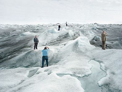 Eine Gruppe von Touristinnen und Touristen steht auf einer schmelzenden Eislandschaft und posiert an verschiedenen Punkten des Bildes für Fotos. An vielen Stellen der Eislandschaft hat sich schwarzer Ruß abgesetzt, das Eis ist dort sehr dunkel. Im Vordergrund ist eine große Pfütze aus Schmelzwasser.