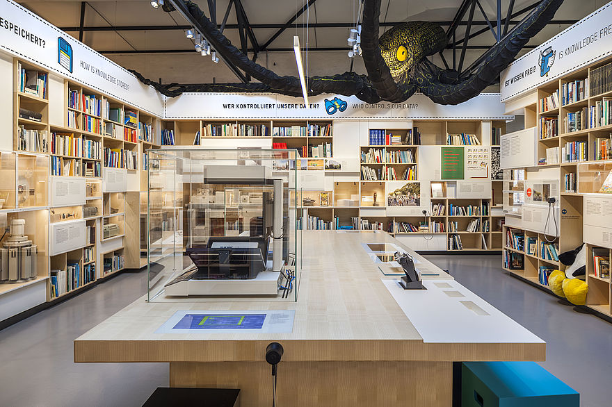 Der Ausstellungsbereich INFORMATION ist wie eine Bibliothek gestaltet. In den vier Wänden sind Regale mit Büchern eingelassen. Im Zentrum steht ein großer Tisch. Darüber schwebt ein riesiger Kraken.