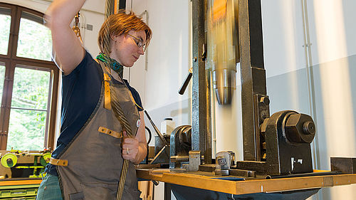 Eine Frau arbeitet an einem Fallhammer. Sie trägt eine Schürze und hält in beiden Händen ein Seil. Der Hammer stürzt herab.