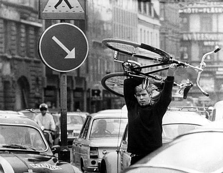 Ein Mann trägt ein Fahrrad über seinem Kopf. Er läuft zwischen im Stau stehenden Autos hindurch. Das Schwarz-Weiß-Foto wurde am 3.09.1967 in Malmö aufgenommen. An diesem Tag stellte Schweden von Links- auf Rechtsverkehr um. In Teilen des Landes führte das zunächst zu Problemen und Staus.