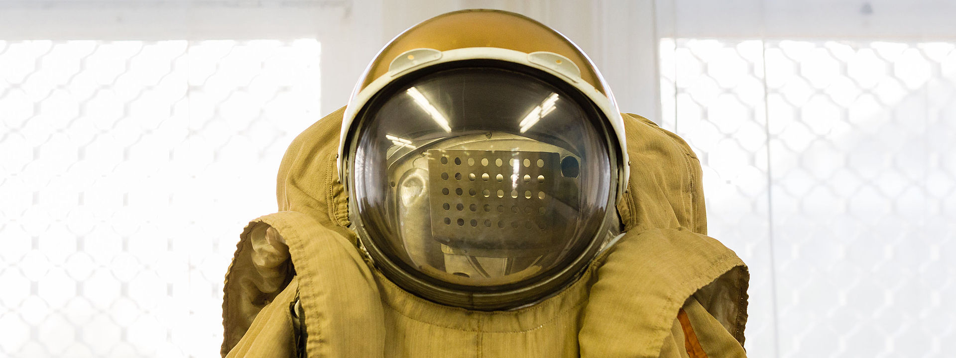 This image is a central view of a cosmonaut’s suit from the waist upwards and includes a spherical helmet with a large glass visor. In the background, an ornamental hanging with a gold fringe is suspended above a window.