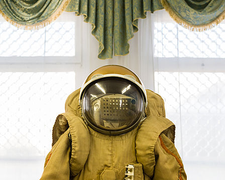Das Bild zeigt zentral einen Kosmonautenanzug von der Taille aufwärts. Darauf sitzt ein kugelförmiger Helm mit großem Glasvisier. Im Hintergrund hängt über einem Fenster eine Zierschabracke mit Goldbordüre.