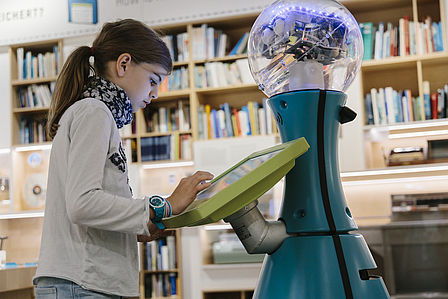 Ein Mädchen tippt auf einen Screen, der an einem humanoiden türkisfarbenen Roboter befestigt ist. Im Hintergrund sieht man einen Ausstellungsraum.