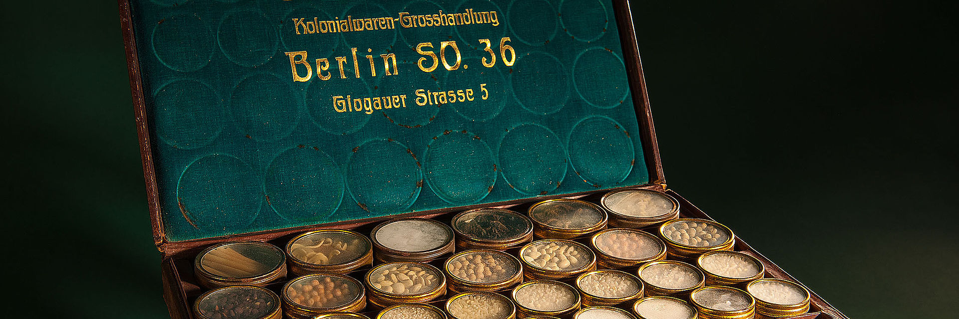Lederkoffer mit verschiedenen kleinen Gefäßen, in denen sich Proben kolonialer Produkte wie Kakao befinden. Die Innenseite des Kofferdeckels trägt den Firmennamen Skibbe & Zorn.