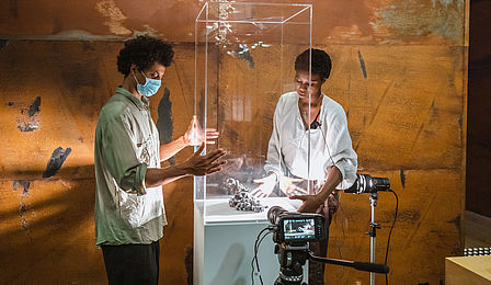 Ein Mann und eine Frau mit dunkler Hautfarbe stehen an einer Glasvitrine, in der ein Haufen Staub liegt. Im Hintergrund ist eine braune Metallwand zu sehen.