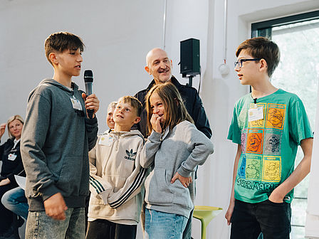 Eine Gruppe von Schülern spricht miteinander, ein Junge hält ein Mikrofon in der Hand, im Hintergrund ist ein Mann zu sehen, der den Kindern zuhört.