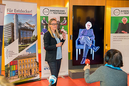 Eine Frau spricht auf einer Presseveranstaltung. Hinter ihr befindet sich ein Bildschirm, der eine ältere Frau zeigt, die auf einem Stuhl sitzt.