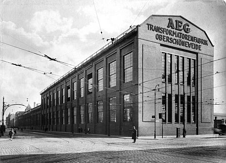 Schwarzweiß-Foto eines großen Fabrikhallengebäudes mit großen Fenstern und einem gewölbten Dach. An der Fassade ist der Schriftzug AEG zu lesen.