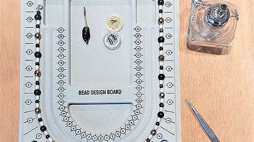 Auf einem Tisch ein Arbeitsbrett zum Knüpfen von Perlenketten, daneben zwei Pinzetten und ein Glasfläschchen.