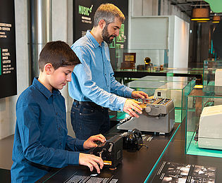 Ein Mann und ein Junge stehen nebeneinander an einem Ausstellungstisch und bedienen eine Schreibmaschine und die Drehscheibe eines Telefons.
